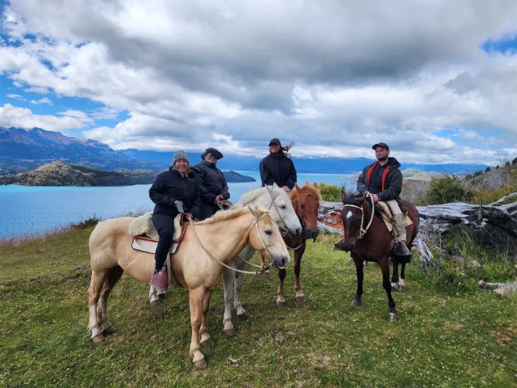 A group of four travelers experiencing horseback riding Patagonia trails on a grassy hilltop, overlooking a beautiful turquoise lake and rugged mountains.