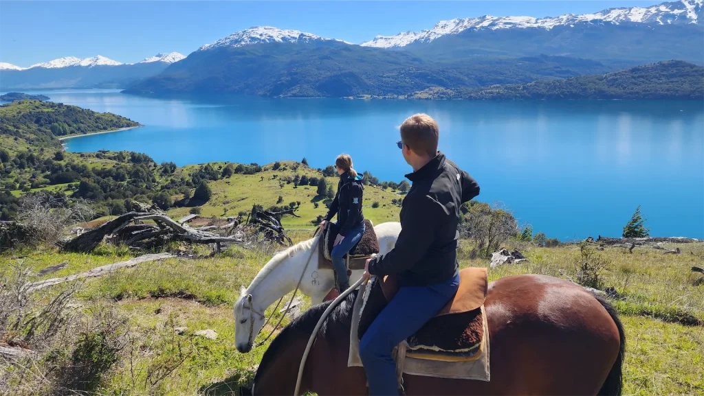 Dos jinetes contemplando un inmenso lago general carrera y montañas nevadas desde una colina verde durante sus excursiones a la patagonia chilena.