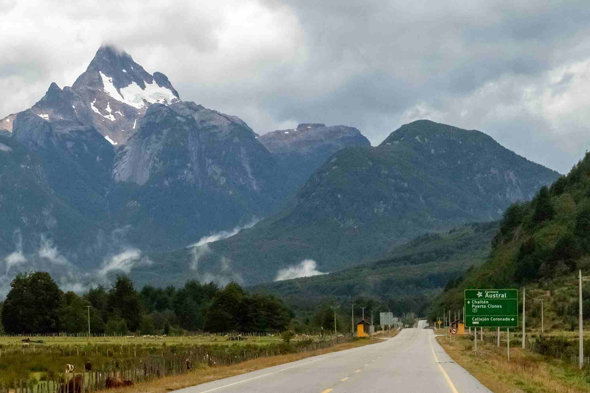 Vista de un tramo pavimentado de la carretera austral bajo un cielo nublado, con imponentes cumbres nevadas de fondo y un letrero vial verde que indica la dirección hacia Chaitén y Puerto Cisnes.