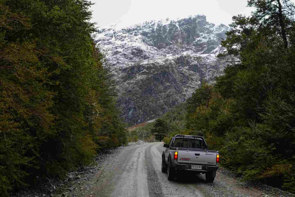 A 4x4 pickup truck navigating the unpaved gravel roads of the carretera austral chilena, surrounded by lush green forests and an imposing snow-capped mountain.