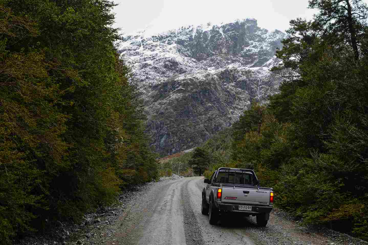 A 4x4 pickup truck navigating the unpaved gravel roads of the carretera austral chilena, surrounded by lush green forests and an imposing snow-capped mountain.